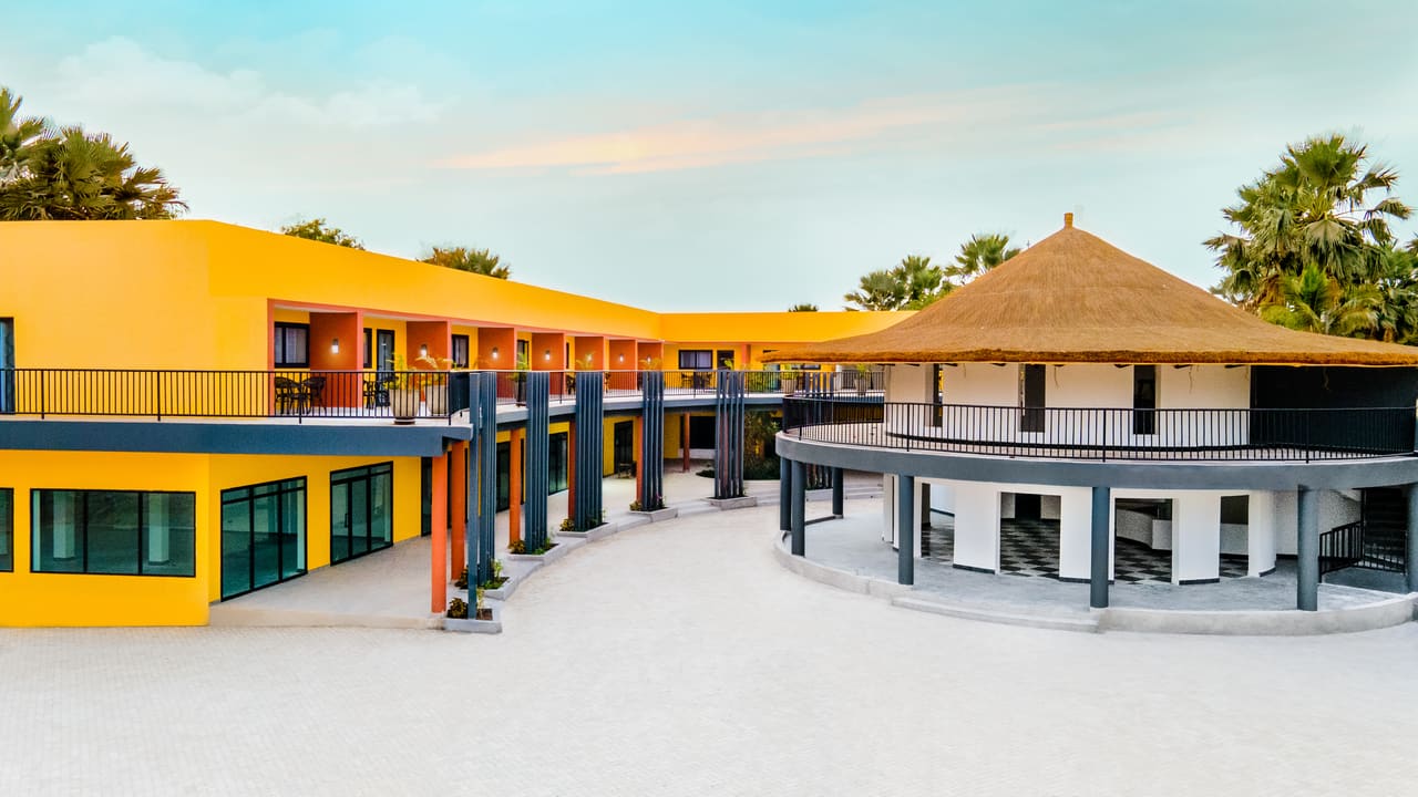 Bakadaji Hotel at dusk: a yellow and orange two-storey guest wing with balconies beside a circular thatched-roof roundhouse