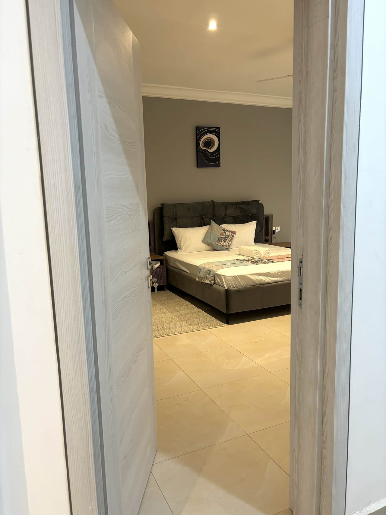 Bedroom in a Fajara Lux apartment seen through the doorway, with upholstered bed and tiled floor