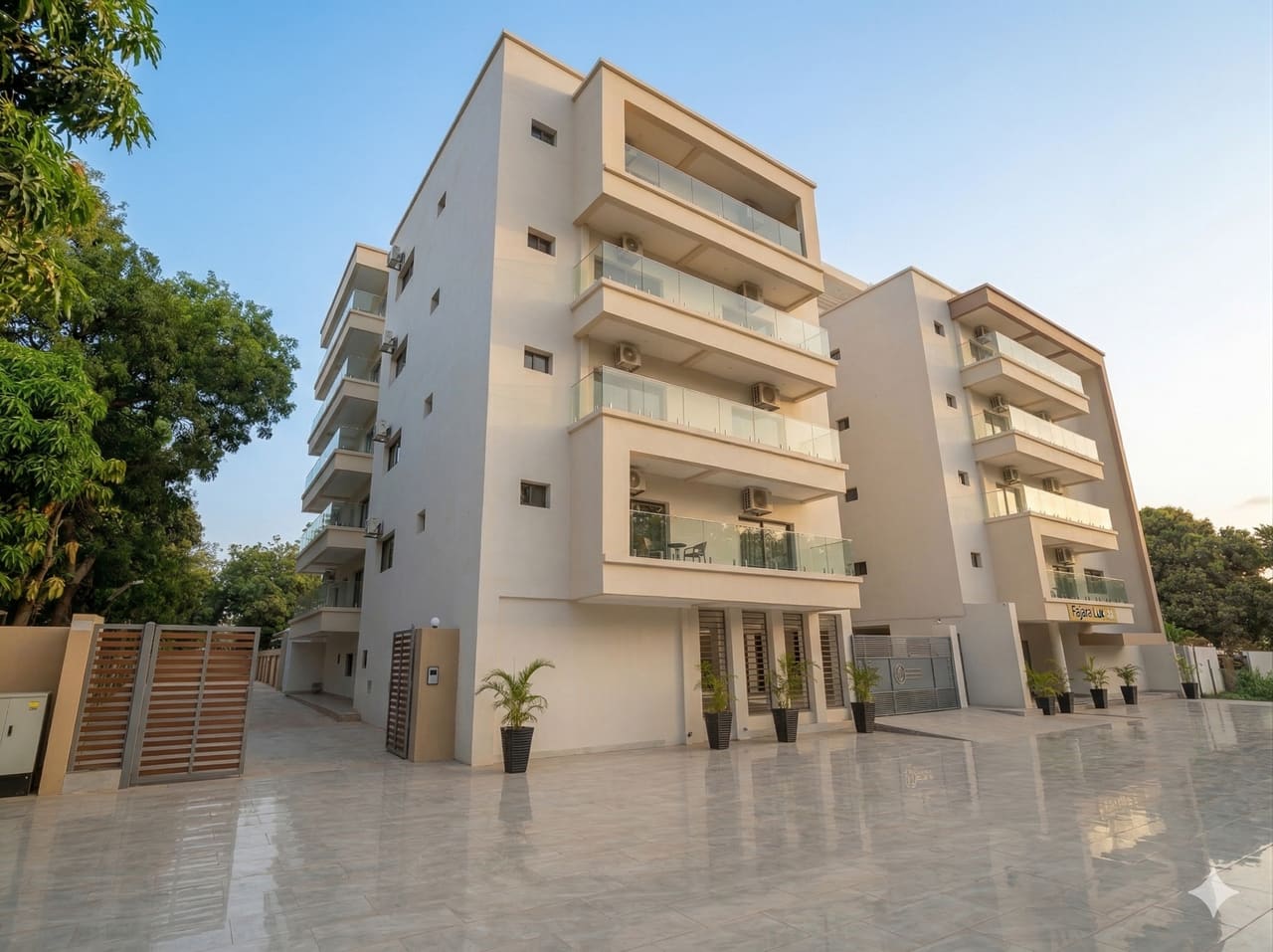 Fajara Lux Twin Homes: a four-storey sand-coloured apartment block with recessed glass-balustrade balconies above a tiled forecourt